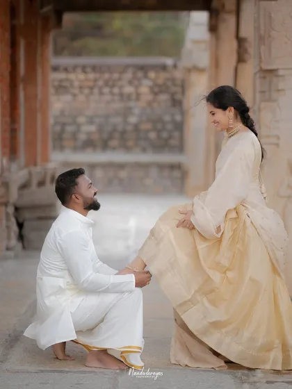 A playful and charming moment as the groom-to-be helps his partner with her footwear inside a temple corridor.
