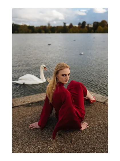 An environmental fashion portrait taken by a lake, featuring a model in a striking red dress alongside a swan. The image combines fashion with nature to create a serene yet dramatic narrative.