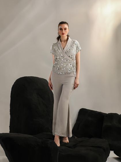 A full-length look featuring an embellished wrap top and flared trousers. The model stands atop black geometric blocks, creating a strong, architectural composition.