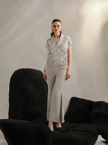 A full-length look featuring an embellished wrap top and flared trousers. The model stands atop black geometric blocks, creating a strong, architectural composition.