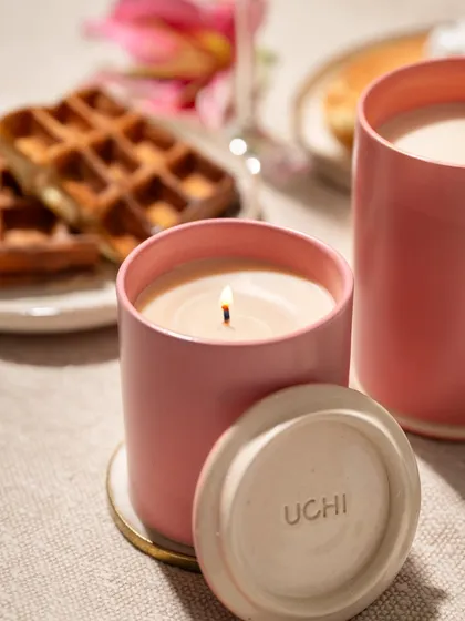 A close-up shot focusing on the warm glow of the candle flame. The blurred background with waffles and flowers adds to the cozy ambiance without distracting from the product itself.