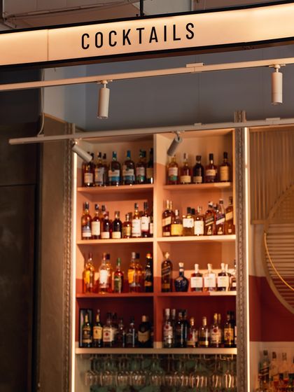 Behind the bar, our shelves are stocked and ready. The "COCKTAILS" sign points to where our mixologists work their magic, using a vast library of spirits.