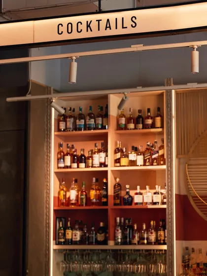 Behind the bar, our shelves are stocked and ready. The "COCKTAILS" sign points to where our mixologists work their magic, using a vast library of spirits.