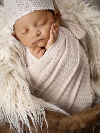 A close-up of a sleeping newborn's face, cradled by their own hands. The soft, fluffy textures of the hat and basket lining create a feeling of ultimate comfort.