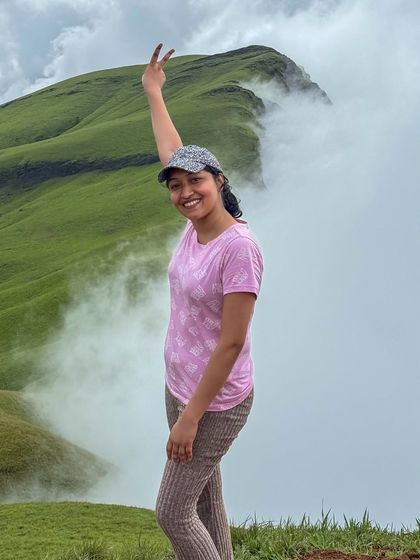 A happy trekker flashing a peace sign with the misty green peaks of Netravathi behind her. The joy of reaching the top is contagious.