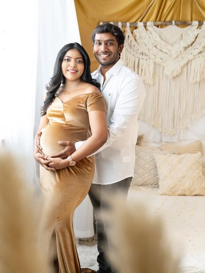 A happy couple posing in a warm, boho-inspired studio. The partner stands behind the expecting mother, both holding her baby bump, sharing a smile in their coordinated gold and white outfits.
