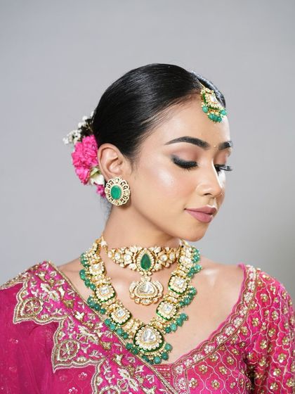 This soft glam bridal look features a simple, elegant bun adorned with a single pink flower. It's a minimalist yet beautiful style that complements the intricate jewelry and makeup, perfect for a modern bride.