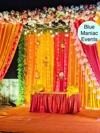 A beautiful Mehendi or Sangeet stage with a backdrop of orange and pink drapes, fairy lights, and a floral border. This setup is perfect for evening celebrations.