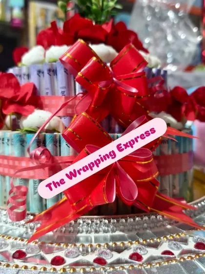 This two-tier cash cake is decorated with bright red ribbons and flowers, sitting on a mirrored platter. It's a popular choice for wedding gift presentations.