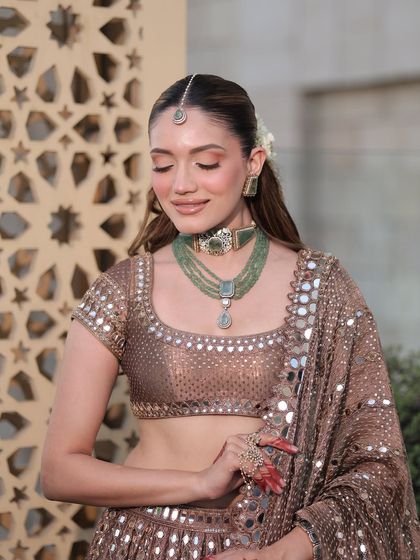 This image showcases the beautiful contrast between the earthy brown lehenga and the vibrant green jewelry, a perfect combination for a daytime event.