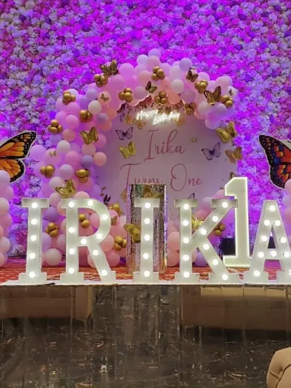 A glamorous butterfly first birthday for Irikia. The stage features a wall of flowers, a balloon arrangement with gold butterflies, and the birthday girl's name in large, bright marquee letters.