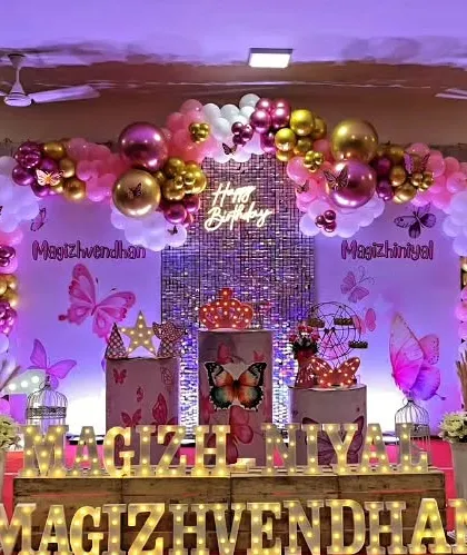 A butterfly theme party on a stage, with a sequin backdrop, neon sign, and a pink, purple, and gold balloon arch.