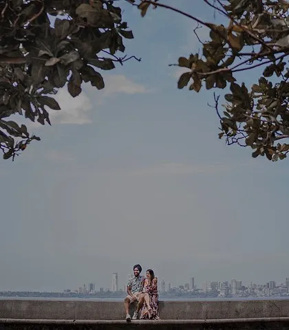 A wide, cinematic shot of Ashleen and Ranjodh sitting together on the Marine Drive promenade, with the famous Queen's Necklace skyline of Mumbai stretching out behind them.