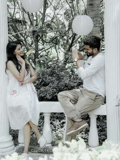 A playful role-reversal with the groom pretending to take a photo of the bride, who strikes a pose in a garden gazebo.