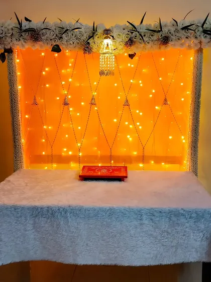 A simple yet elegant setup for a home puja or festival, with a table covered in white fur cloth against a lit-up backdrop.