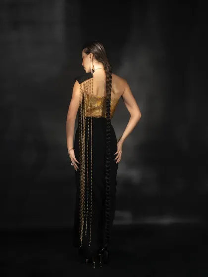 A view from the back of the black and gold Nikhil Thampi outfit. This shot is designed to highlight the dramatic, floor-length chain detailing and the unique braid hairstyle.