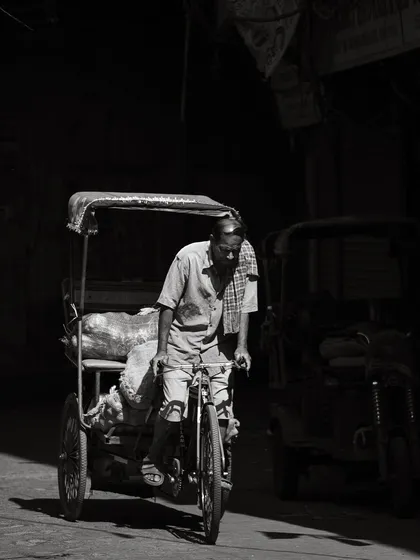 A cycle-rickshaw driver pedals through a dark lane, his form highlighted by a sliver of light. This black and white image captures the daily toil and quiet dignity of workers in Old Delhi.