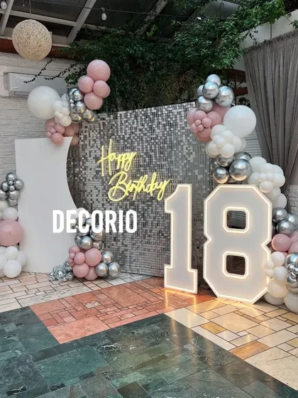 An exciting 18th birthday celebration with a glamorous silver sequin wall that creates a stunning photo opportunity. The setup includes large, illuminated '18' marquee numbers, a 'Happy Birthday' neon sign, and a balloon arch in shades of pink, silver, and white.