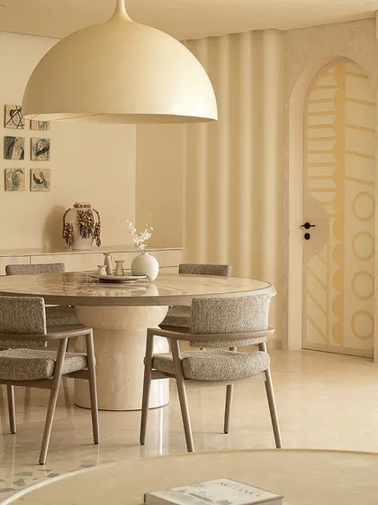 A cozy corner in the 'Soft Serve' apartment, featuring a round dining table under a large dome light. The space is designed with soft curves and a neutral palette to evoke a sense of calm and tranquility.