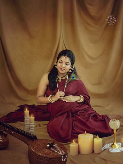 A serene portrait of a mom-to-be in a deep red saree, surrounded by candles and a traditional veena, creating a musical and spiritual ambiance.