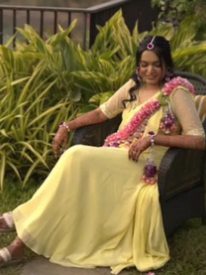 A relaxed moment during a Haldi celebration. The bride is wearing a long floral garland made of pink and purple flowers, which adds a pop of colour to her yellow outfit.