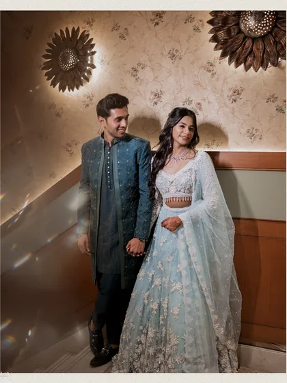 A stylish full-length portrait of the couple against a decorated wall. The warm lighting from the sunflower-shaped sconces adds a beautiful ambiance to this elegant engagement photo.