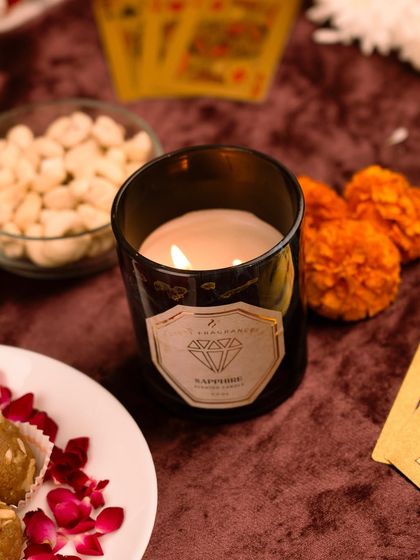 A scented candle in a dark glass jar, styled with marigold flowers and other festive items. The composition is warm and inviting.