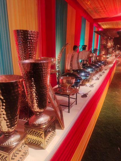 A long shot of a buffet line at a colorful, themed event. The setup features hammered copper vases and stainless steel chafing dishes against a vibrant red, yellow, and blue backdrop.