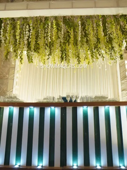 A stylish bar setup for a daytime event. The backlit green and white striped bar is topped with a hanging installation of lush greenery, creating a fresh and modern look.