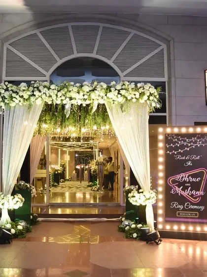 A glamorous entrance for a ring ceremony, featuring a marquee-style welcome sign and an archway draped in white fabric and florals, illuminated for a nighttime event.