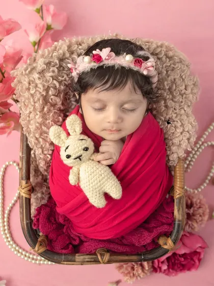 A newborn baby girl wrapped in a vibrant red swaddle, holding a tiny bunny. The contrast of the red against the soft pink background creates a striking and beautiful image.