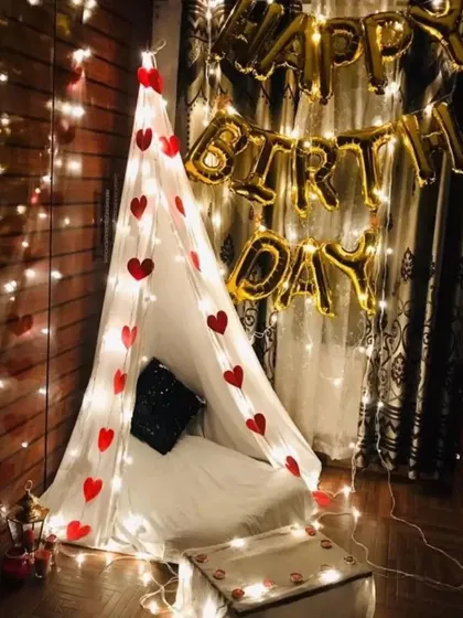 A cozy and intimate birthday celebration created with a teepee tent. The tent is decorated with fairy lights and red hearts, with 'Happy Birthday' foil balloons in the background.