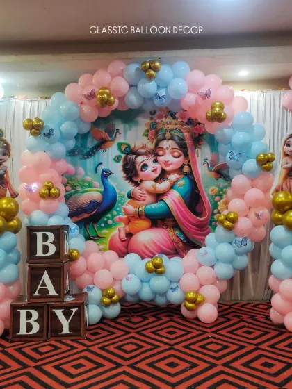 A traditional Radha Krishna theme for a baby shower. This setup includes a custom printed backdrop, character cutouts, and a circular balloon arch in soft pink and blue, accented with gold balloons and wooden 'BABY' blocks.