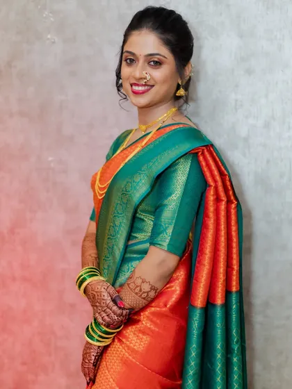 A happy portrait from an engagement ceremony. The bride-to-be looks radiant in a vibrant orange and green silk saree, her hands adorned with henna and traditional green bangles.