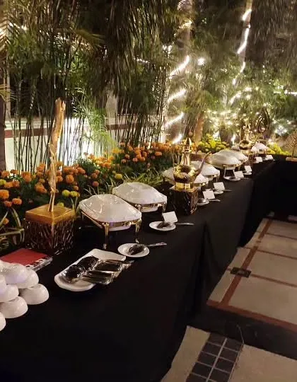 A night-time buffet set amidst lush palm trees and fairy lights. The table is dressed in black, with white serving dishes and floral arrangements of marigolds creating a beautiful contrast.