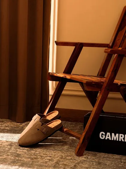 An atmospheric shot of clogs next to a wooden chair and dark curtain. The play of light and shadow adds a touch of sophistication and warmth to this simple footwear style.