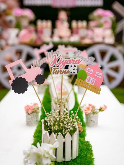 The kids' table is set for fun with a faux grass runner and custom centerpieces. Each centerpiece features themed cutouts like a pink tractor and barn, adding a playful touch to the party decor.