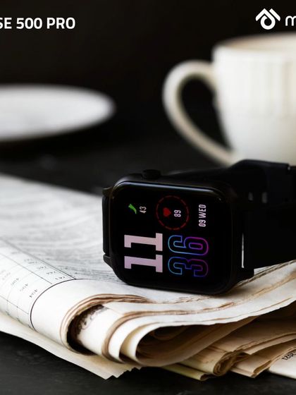The smartwatch is placed on a folded newspaper next to a coffee cup. This classic morning routine setup gives the modern tech a timeless, relaxed, and sophisticated feel.