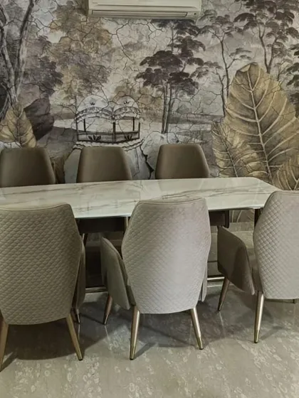 A grand dining set featuring a long, white marble table and eight quilted, upholstered chairs. The artistic wallpaper in the background complements the furniture, creating a dining room that is both elegant and expressive.