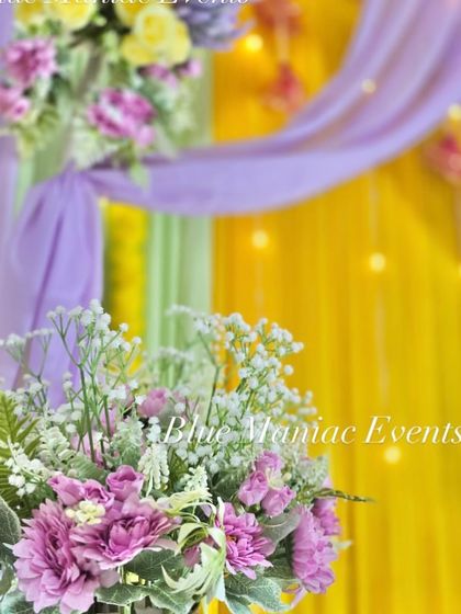 A close-up shot of a Haldi setup, focusing on the beautiful artificial flower arrangements in shades of purple and white, set against a lavender and yellow backdrop.
