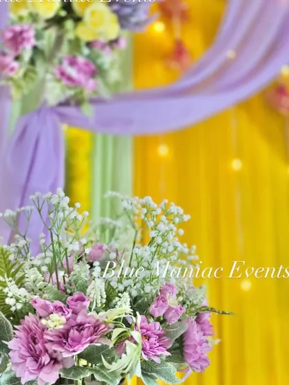 A close-up shot of a Haldi setup, focusing on the beautiful artificial flower arrangements in shades of purple and white, set against a lavender and yellow backdrop.