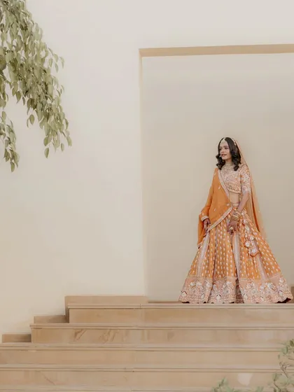 A full-length bridal portrait showcasing the magnificent orange lehenga against a minimalist architectural backdrop. This shot is all about grace and style.