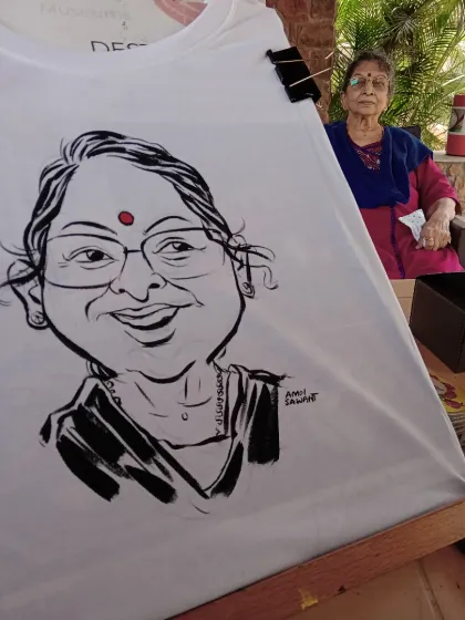 A freshly painted caricature on a t-shirt, with the smiling subject sitting right behind it. I paint these live for guests to watch.