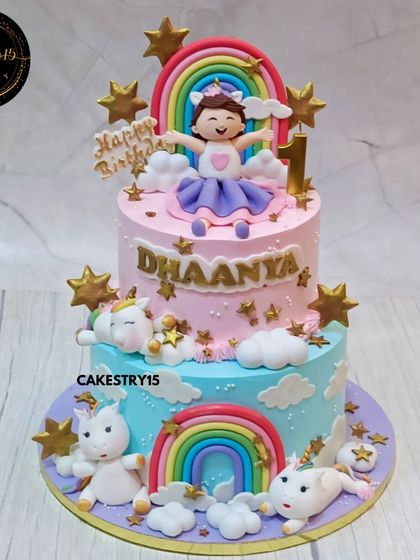A dazzling two-tier rainbow unicorn cake for a first birthday. This cake, in pineapple and black forest flavors, is adorned with fondant unicorns, rainbows, clouds, and golden stars.