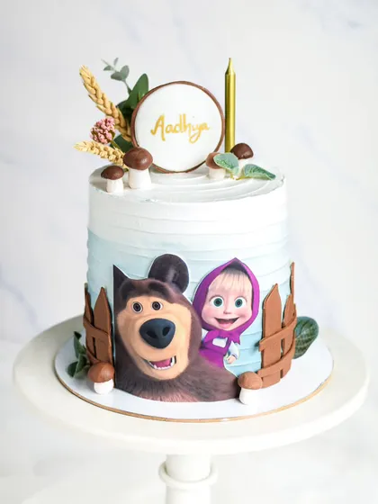 A cute Masha and the Bear themed cake. The design uses an edible print of the characters and is decorated with rustic elements like a wooden fence, mushrooms, and wheat stalks.