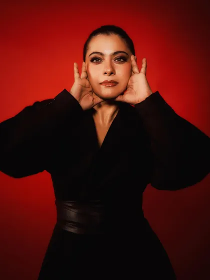 Kajol in a dramatic all-black wrap dress with a leather belt, set against a bold red background for the 'Do Patti' promotions.