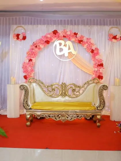 An elegant engagement stage with a pink floral ring, a custom 'BA' monogram, and a beautiful gold and yellow diwan.