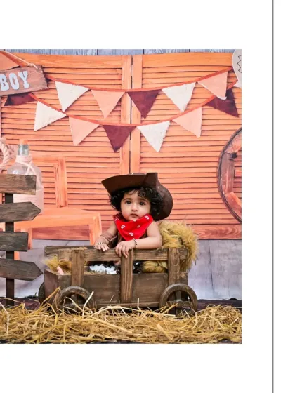 Seated in a wooden cart, this little cowboy looks ready to round up some cattle. The rustic barn-door background and hay-strewn floor complete this adorable western theme.