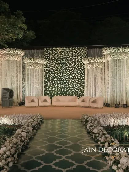 A breathtaking reception stage designed to look like a palace of flowers. The pathway is lined with blooms, leading to a seating area set against a wall entirely made of white flowers, creating a truly immersive experience.