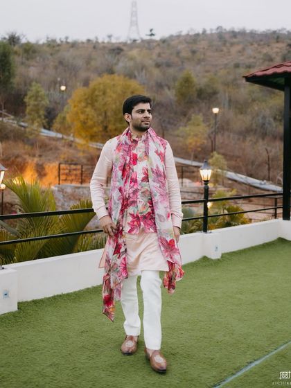 A full-length shot of the groom's mehendi outfit. The floral jacket and scarf paired with a simple kurta and white pants create a look that is stylish, comfortable, and perfect for a sundowner event.